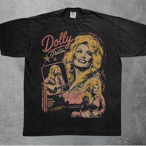 May include: A black short-sleeved t-shirt featuring a graphic print of Dolly Parton. The design includes the text Dolly Parton, the phrase what would dolly do?, and three images of the singer. The print uses muted yellow, pink, and white colours.