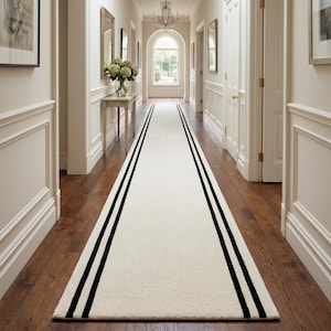 Peut inclure: Un long tapis de couloir rectangulaire présentant un centre de couleur crème uni, bordé de deux bandes noires parallèles de chaque côté. Le tapis est placé au centre sur un plancher en bois sombre, s'étendant le long d'un couloir lumineux.