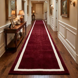 May include: A long, rectangular runner rug featuring a deep burgundy red center and a thick, contrasting cream-colored border. The rug is positioned centrally on a dark hardwood floor, extending down the length of a hallway with white walls.