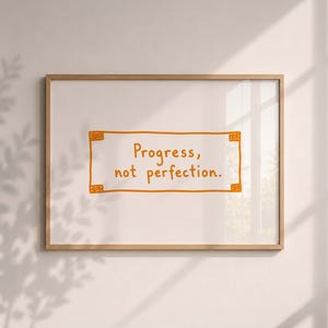 May include: A rectangular wooden frame containing a white print with an orange hand-drawn border and the text "Progress, not perfection." in orange lettering. The frame is displayed against a neutral wall with soft, natural light and shadows.
