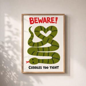 Op de afbeelding: Een ingelijste kunstdruk op een witte achtergrond met een groene slang met donkere strepen die in een hartvorm is gekronkeld. De tekst BEWARE! staat in het rood bovenaan en de tekst CUDDLES TOO TIGHT staat in het zwart onderaan.