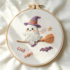 May include: A circular wooden embroidery hoop containing white fabric with a hand-stitched design of a white ghost wearing a purple witch hat, riding a broomstick, accompanied by a small star, a purple bat, and two striped candies.