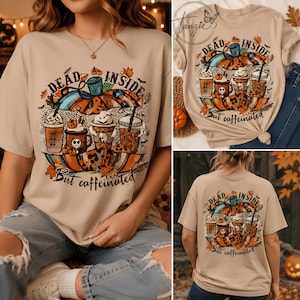 May include: A beige short-sleeve t-shirt featuring a graphic of a large pumpkin and four coffee cups. The text reads "DEAD INSIDE" at the top and "But caffeinated" at the bottom. The design includes autumn leaves, small bats, and leopard print accents.