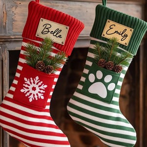 Op de afbeelding: Twee gebreide kerstsokken hangen aan een open haard. De ene is rood-wit gestreept met een witte sneeuwvlok en een naamplaatje Bella. De andere is groen-wit gestreept met een witte pootafdruk en een naamplaatje Emily.