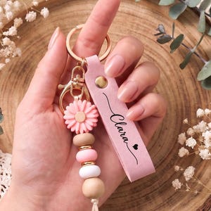 以下が含まれることがあります： 「Clara」という名前と小さなハートがプリントされたピンクのレザーストラップが付いたキーホルダー。ゴールドトーンの金属リングに、ピンクのフラワービーズ、2つの丸いピンクのビーズ、1つの白いビーズ、1つの天然木のビーズが連なったチャームが取り付けられています。