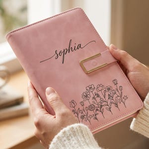 Peut inclure: Un carnet rose clair doté d'un fermoir magnétique rectangulaire doré. La couverture est personnalisée avec le prénom "sophia" écrit en lettres cursives noires et comporte une illustration au trait noir de fleurs sauvages en bas.