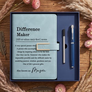 May include: A light blue notebook with a magnetic closure and a matching blue pen in a gift box. The cover features the printed definition of Difference Maker and the name Mason. The notebook and pen are set against a wooden background.