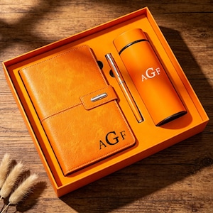 May include: An orange gift set in a matching box containing a textured orange notebook with a metal clasp, a slim orange pen, and an orange cylindrical thermos. Both the notebook and the thermos are personalised with the black monogrammed letters AGF.