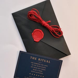 Puede incluir: Un sobre negro sellado con un sello de cera roja con un diseño de árbol, coronado con un cordel rojo enrollado. Debajo, una tarjeta rectangular azul oscuro muestra el texto: THE RITUAL, ASK SOMEONE YOU LOVE TO TIE SEVEN KNOTS.