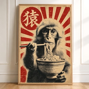 以下が含まれることがあります： 箸を使ってボウルから麺を食べている猿の白黒イラストが描かれた額入りアートプリント。背景には、赤い太陽のグラフィックと放射状のストライプ、そして猿を意味する日本語の漢字がデザインされています。