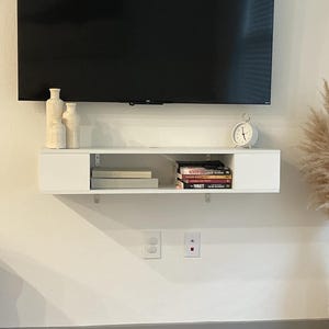 May include: A wall-mounted white floating shelf positioned beneath a large black television. The shelf features two closed side compartments and an open center section holding stacked books. Three beige vases and a white analog clock sit on top.