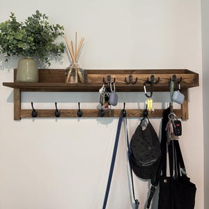 May include: A dark brown wooden wall-mounted shelf featuring a top ledge and two rows of black metal hooks. The shelf holds a green potted plant and a glass reed diffuser. Various items including keys, a blue bag, a black cap, and a black tote hang below.