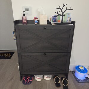 May include: A dark gray wooden shoe storage cabinet featuring two tilt-out drawers with a prominent X-shaped cross-brace design on each front panel. The cabinet has a flat top surface and small round black knobs for opening the storage compartments.