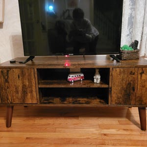May include: A rectangular media console with a dark brown wood grain finish, featuring two side cabinets with doors and two open central shelves. The unit is supported by four tapered legs and holds a flat-screen television on its top surface.