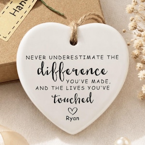 以下が含まれることがあります： 白いハート型のセラミック製オーナメント。黒い文字で「NEVER UNDERESTIMATE THE difference YOU'VE MADE, AND THE LIVES YOU'VE touched」と書かれ、その下に小さなハートのアイコンと「Ryan」という名前が記されており、茶色の麻紐のループで吊り下げられています。