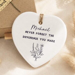 以下が含まれることがあります： Michael NEVER FORGET THE DIFFERENCE YOU MAKEという文字と花のイラストがプリントされた白いハート型のセラミック製オーナメント。Hand madeのラベルと黄色い花が添えられた茶色の段ボール製ギフトボックスが付属しています。