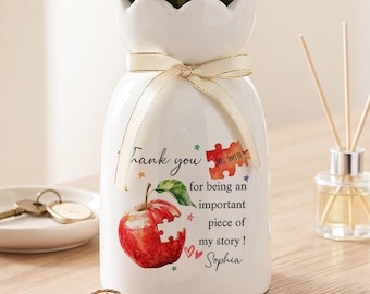 Könnte beinhalten: Eine weiße Keramikvase mit gewelltem Rand, gebunden mit einem cremefarbenen Band, gefüllt mit grünem Blattwerk und weißen Blüten. Die Vase zeigt eine rote Apfelgrafik mit einem fehlenden Puzzleteil und den Text: Thank you for being an important piece of my story! Mrs. Smith, Sophia.