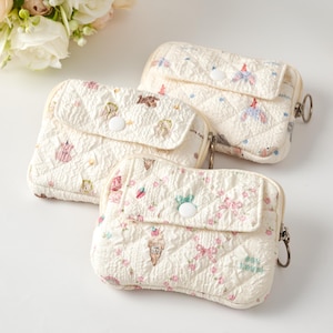 May include: Three small, rectangular, quilted pouches in off-white fabric. Each pouch features a front flap with a white snap button closure, a side zipper with a metal ring pull, and various small, colorful printed motifs including bows and ice cream.