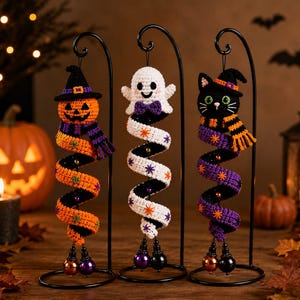 May include: Three crocheted Halloween ornaments hanging from black metal stands. The left is a pumpkin with a hat and scarf, the center is a white ghost with a purple bow, and the right is a black cat with a hat and striped scarf.