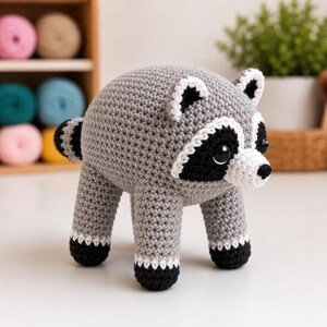May include: A crocheted raccoon plush toy featuring a round grey body, a white muzzle, black eye patches, and black-tipped paws. The creature has small pointed ears with white trim and a striped tail, crafted with visible yarn stitches.