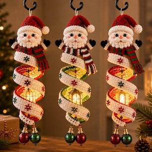 Könnte beinhalten: Drei hängende Weihnachtsanhänger mit gehäkelten Weihnachtsmannfiguren, roten Mützen und gestreiften Schals. Jede Figur sitzt auf einem weißen Spiralband mit roten und grünen Schneeflockenmustern, das eine kleine leuchtende Laterne umschließt.