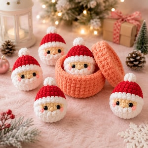 Puede incluir: Un conjunto de cinco pequeñas figuras de Papá Noel tejidas a ganchillo, redondas, con gorros rojos, pompones blancos y barbas blancas, junto a un recipiente de ganchillo color melocotón con tapa, todos con ojos de cuentas negras y mejillas rosas.