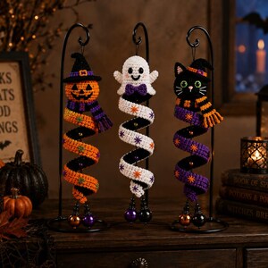 May include: Three crocheted Halloween ornaments hanging from black metal stands. The left is a pumpkin with a witch hat and scarf, the center is a white ghost with a purple bow tie, and the right is a black cat with a witch hat and striped scarf.
