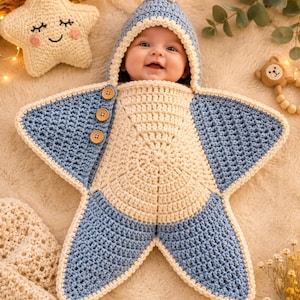 Op de afbeelding: Een stervormige gehaakte baby-inbakerdeken in een lichtblauw en crèmekleurig schema. Het kledingstuk heeft een capuchon met een klein sterretje bovenop, drie houten knopen aan de zijkant en een centraal cirkelvormig patroon in crèmekleurig garen.