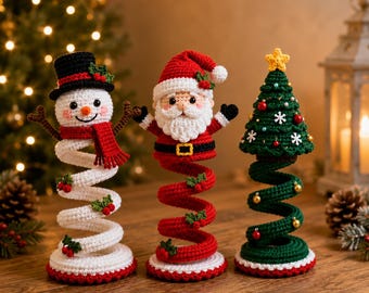 Op de afbeelding: Drie gehaakte kerstversieringen bestaande uit een sneeuwpop met een zwarte hoge hoed en rode sjaal, een kerstman met een witte baard en rode muts, en een groene kerstboom met een gele ster, allemaal op flexibele, spiraalvormige onderstellen.