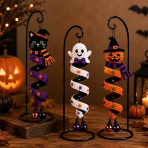 May include: Three crocheted Halloween ornaments hanging from black metal stands. The ornaments feature a black cat, a white ghost, and an orange pumpkin, each wearing a hat and scarf with a spiral body decorated with small star-shaped accents.
