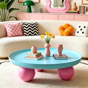 May include: A round light blue coffee table with four large pink spherical legs, positioned on a cream-colored rug with a black wavy border. The table holds a small pink vase, a pink face-shaped vase, books, and a bouquet of yellow daffodils.