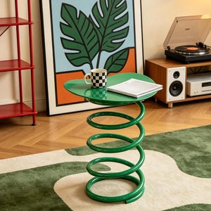 May include: A green side table featuring a flat, heart-shaped top supported by a large, coiled metal spring base. A black and white checkered mug and a closed white book are placed on the table surface, which has a smooth, glossy finish.