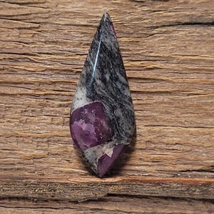 Peut inclure: Une pierre précieuse en forme de goutte d'eau avec une surface polie. La pierre présente une combinaison de couleurs saisissante, notamment le violet profond, le gris et le blanc. La pierre est posée sur un fond en bois rustique.