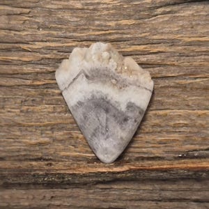 Puede incluir: Una gema pulida en forma de corazón con un aspecto en capas. La piedra presenta bandas de gris y blanco, con una parte superior cristalina. La gema se encuentra sobre un fondo de madera texturizada.
