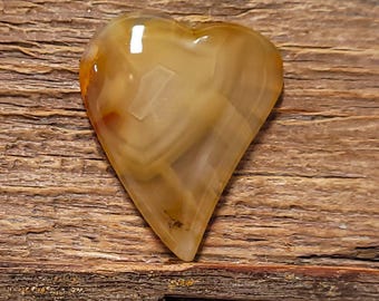 Puede incluir: Una piedra en forma de corazón con una superficie pulida. La piedra presenta bandas de color beige, marrón y crema. El corazón está sobre una superficie de madera envejecida.