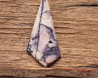 May include: A polished gemstone with a unique, elongated diamond shape. The stone features a blend of white, lavender, and dark gray hues, with intricate veining patterns. The gemstone is set against a rustic wooden background.