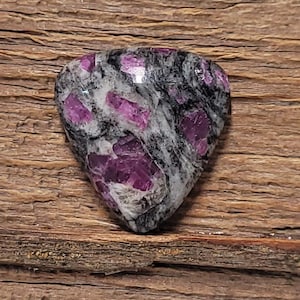 May include: A heart-shaped gemstone with a mix of colors. The stone features a speckled pattern of black, white, and gray, with vibrant magenta-colored inclusions. The gemstone is set against a wooden background.