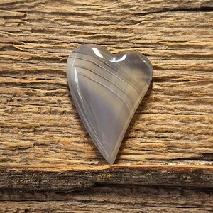 May include: A heart-shaped polished stone featuring smooth, translucent gray surfaces with distinct, thin, parallel white banding lines running diagonally across the face, creating a layered appearance with a glossy, reflective finish.