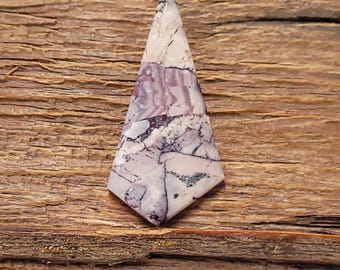 May include: A polished, triangular gemstone with a pointed top. The stone features a mix of white, lavender, and black colors, with intricate veining patterns. The gemstone is set against a rustic, wooden background.