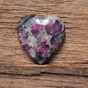 Puede incluir: Una piedra preciosa en forma de corazón con una mezcla de tonos grises, blancos y negros. La piedra presenta vibrantes cristales de rubí rosa intenso incrustados en su superficie. La piedra preciosa se muestra sobre un fondo de madera rústica.
