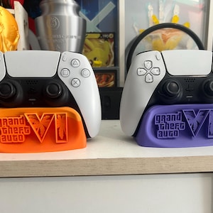 Puede incluir: Dos mandos de juego blancos y negros sobre soportes personalizados. El soporte izquierdo es naranja con el texto Grand Theft Auto VI, y el derecho es morado con el mismo texto. Ambos soportes presentan siluetas de palmeras en el número romano VI.