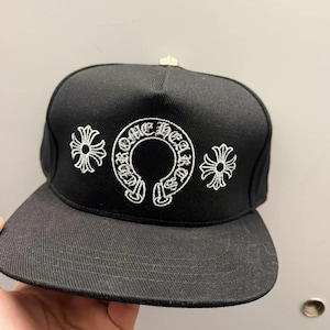 May include: A black baseball cap featuring white embroidered text that reads Chrome Hearts in a circular design. The cap includes two white embroidered cross symbols on either side of the text and a small silver-toned button on the top crown.
