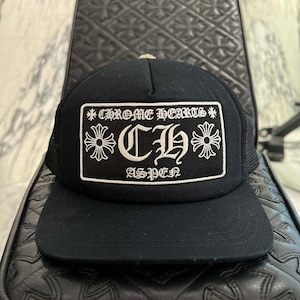 Op de afbeelding: Een zwarte truckerpet met een rechthoekige patch aan de voorzijde. De patch toont de tekst CHROME HEARTS, de letters CH en het woord ASPEN in een gotisch lettertype, geflankeerd door twee kruissymbolen op de voorzijde.