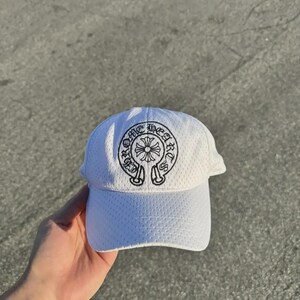 May include: A white baseball cap featuring a textured mesh fabric and a black embroidered circular logo on the front panel. The logo displays a central cross emblem surrounded by the text CHROME HEARTS in a gothic-style font. The cap has a curved brim.