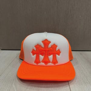 以下が含まれることがあります： 鮮やかなオレンジと白のトラッカーキャップ。フォーム素材のフロントパネルには、オレンジ色の刺繍が施された3つの十字架のエンブレムがあしらわれています。オレンジ色のメッシュバック、ステッチ入りのオレンジ色のつば、そしてトップには小さな銀色のボタンが付いています。