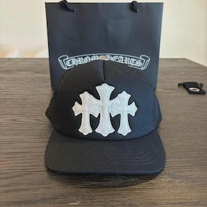 May include: A black baseball cap featuring three white cross-shaped patches on the front panel. The cap has a structured crown and a flat brim. Behind the cap sits a black shopping bag with the white text CHROME HEARTS printed on the front.