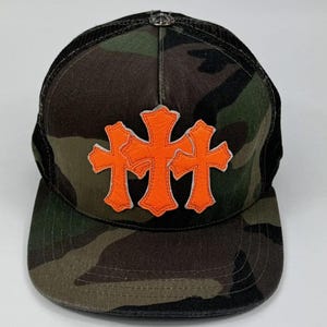 Può includere: Un cappellino trucker con stampa mimetica e tre applicazioni a forma di croce color arancione brillante sul pannello frontale. Il cappello presenta una visiera piatta, una parte posteriore in rete nera e un piccolo bottone metallico in cima.