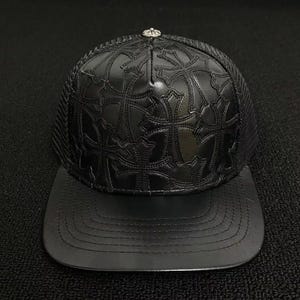 May include: A black trucker hat featuring a front panel adorned with an embossed cross pattern. The hat includes a flat brim, a mesh back section, and a small silver-toned circular button detail at the very top center of the crown structure.