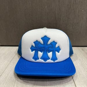 Op de afbeelding: Een truckerpet met een wit schuimrubberen voorpaneel voorzien van drie blauwe kruisapplicaties. De pet heeft een blauwe klep en blauwe mesh zij- en achterpanelen. Een kleine zilverkleurige metalen knoop bevindt zich boven op de kroon.