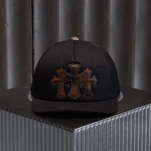 Puede incluir: Una gorra tipo trucker negra con parte trasera de malla y panel frontal sólido. El frente muestra tres apliques de cuero marrón en forma de cruz dispuestos horizontalmente. Un pequeño botón de metal plateado se encuentra en la parte superior.